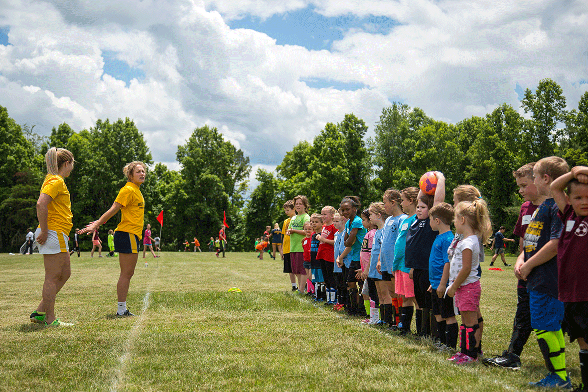 Children stand in a line on the field as WVU women's soccer players 