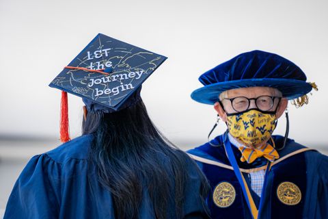 Graduate wearing decorated mortarboard that reads 'Let the Journey Begin' walks by President Gee