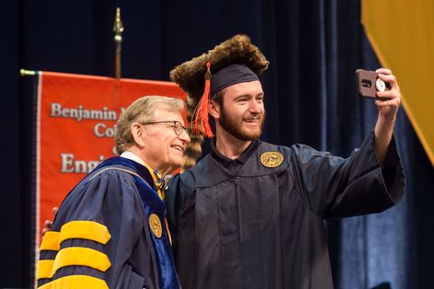 Student dressed in graduation cap and gown takes selfie with President Gee