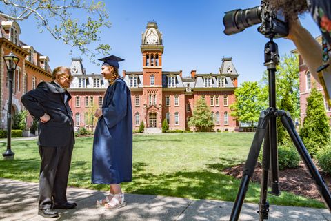 President Gee talks to a student dressed in a graduation cap and gown with Woodburn Hall in the background.